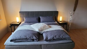 A bed with a grey upholstered headboard, two bedside tables with lamps, and a decorative plant on the right.
