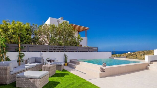 A modern outdoor pool area with a white building, green lawn, and wicker furniture. - Aegean Blue Apartment (Ligaria)