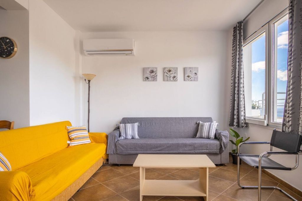 A living room with a yellow sofa, a grey sofa, a wooden coffee table, and a black chair. There are three wall-mounted artworks above the grey sofa.