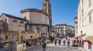 Una plaza histórica con edificios de piedra, una torre destacada y zonas de estar al aire libre.