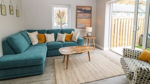A modern living room with a teal sofa, a patterned armchair, a wooden coffee table, and a large window offering a view of the outdoors.