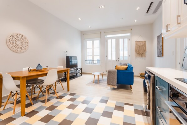 A modern living room with a wooden dining table, white chairs, a blue sofa, and a kitchen area with a washing machine. - Casa Love 1A (Panama City)