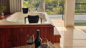 Une pièce avec un jacuzzi, une table en verre avec des verres à vin et une bouteille, et une vue sur une montagne et la verdure.