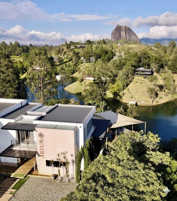 Un edificio moderno con techo plano, placa con el nombre a la vista, y una vista de un lago y zona boscosa al fondo.