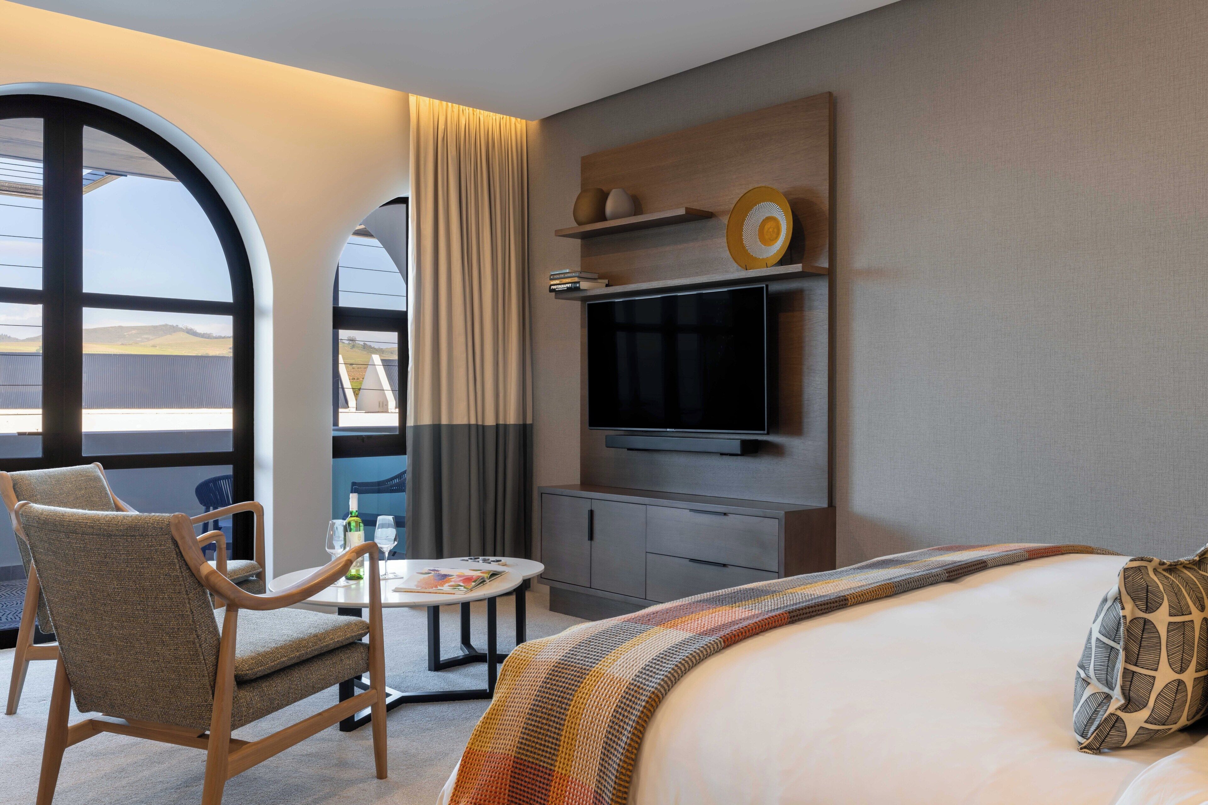 Quarto de hotel com uma cama grande, televisão, uma mesinha com cadeira e vista pela janela.
