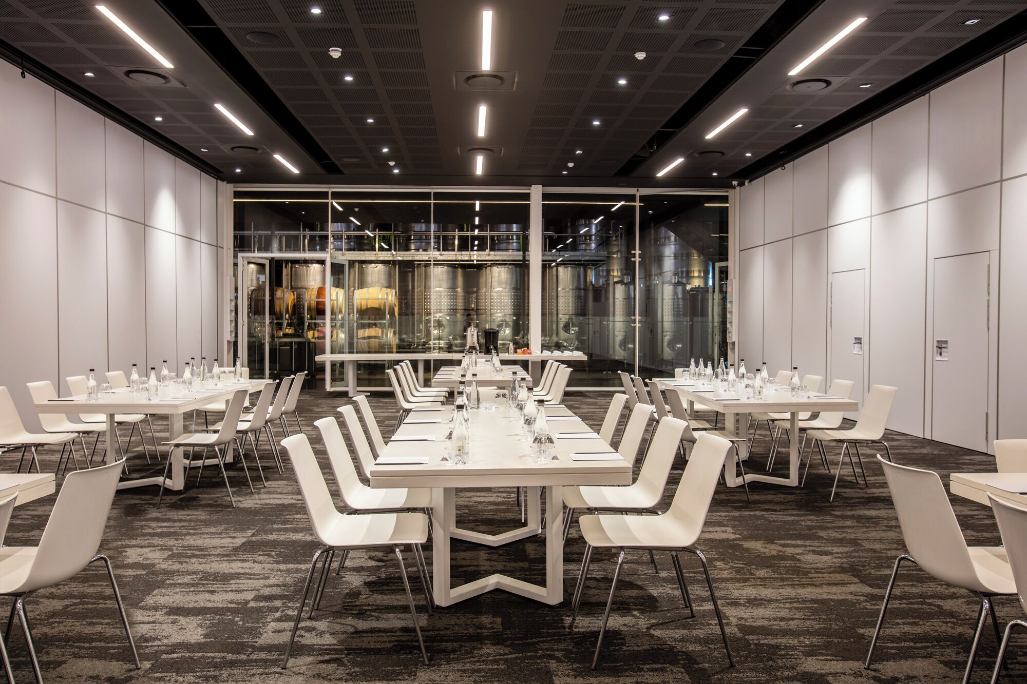A conference room with multiple tables and chairs, a carpeted floor, and a glass wall with industrial elements.