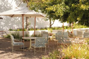 An outdoor seating area with tables and chairs, shaded by umbrellas, surrounded by greenery.