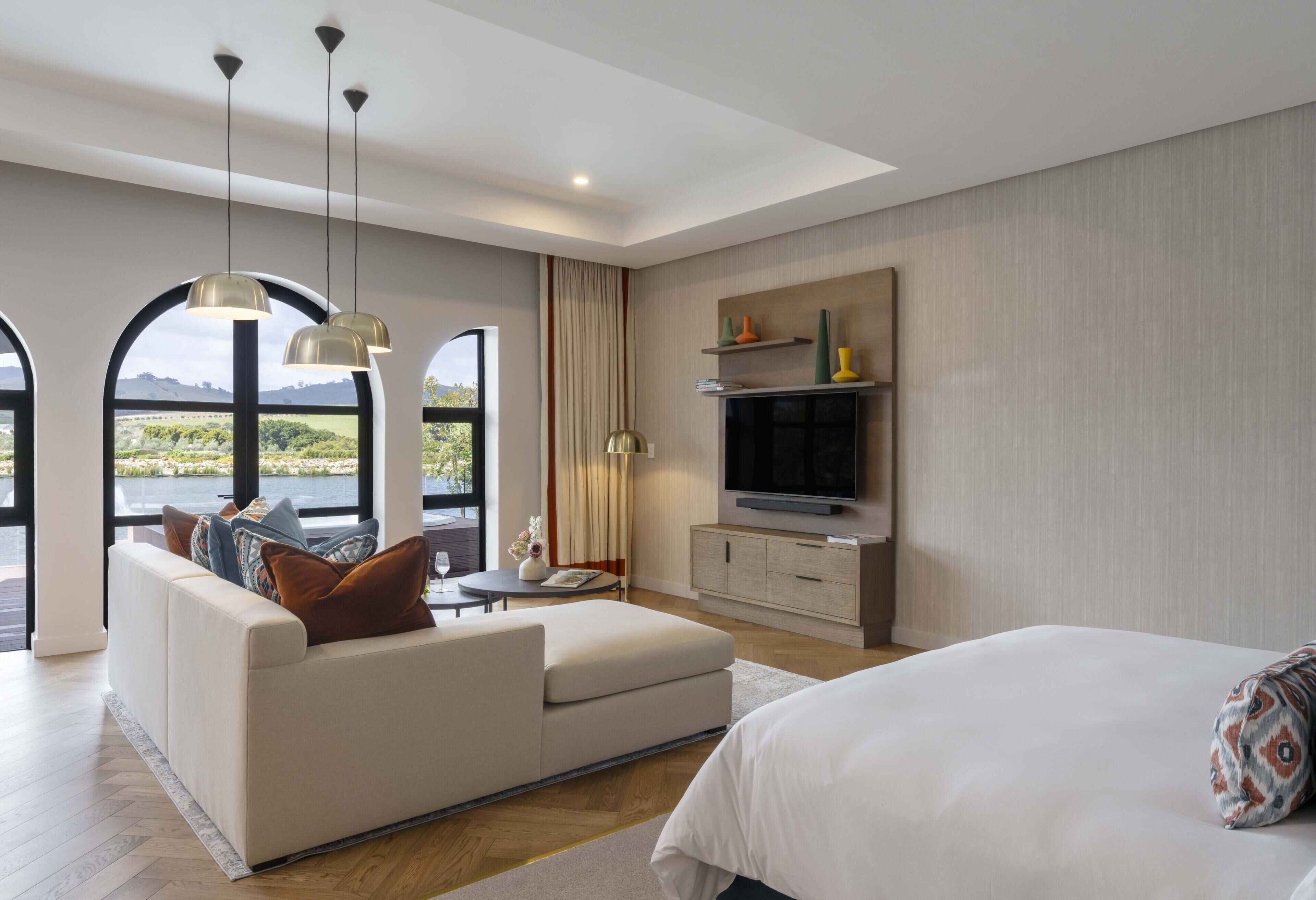 A modern hotel room with a large bed, a sofa, a TV, and a view of a body of water.