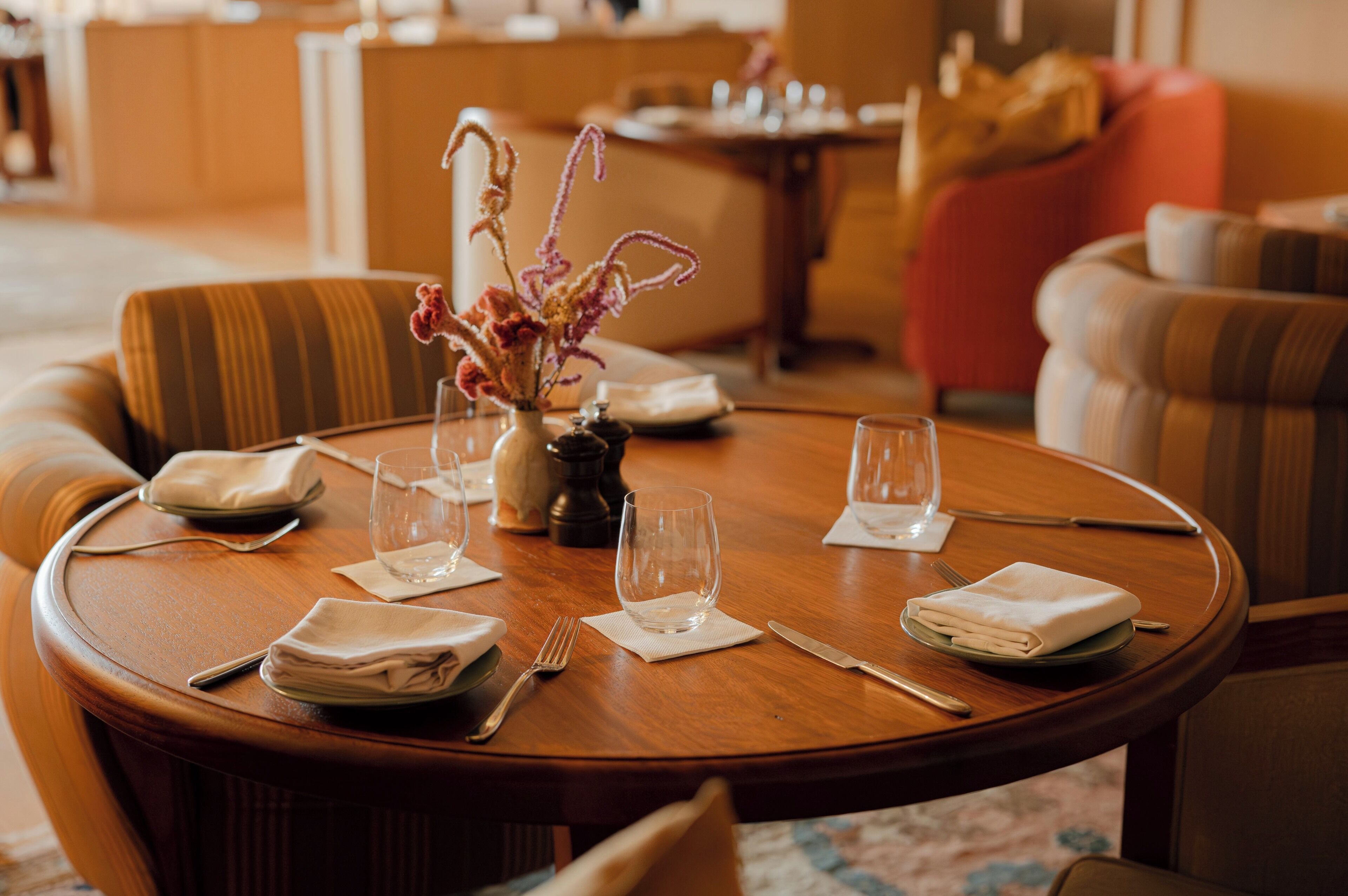 A round dining table set with plates, cutlery, and glasses, surrounded by striped armchairs and a sofa in a well-lit room.