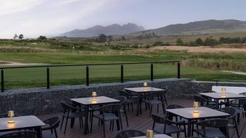 Área externa com mesas e cadeiras, com vista para um campo de golfe e colinas ao longe.