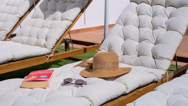 A beachside lounge area with cushioned chairs, a straw hat, sunglasses, and a book.