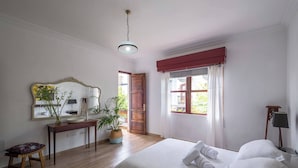 A hotel room with a bed, a desk with a mirror, a small stool, a potted plant, and a window with curtains.