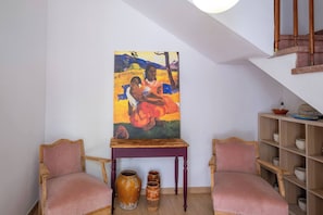 A room with two pink upholstered chairs, a wooden table with a painting, and a shelf with pottery.