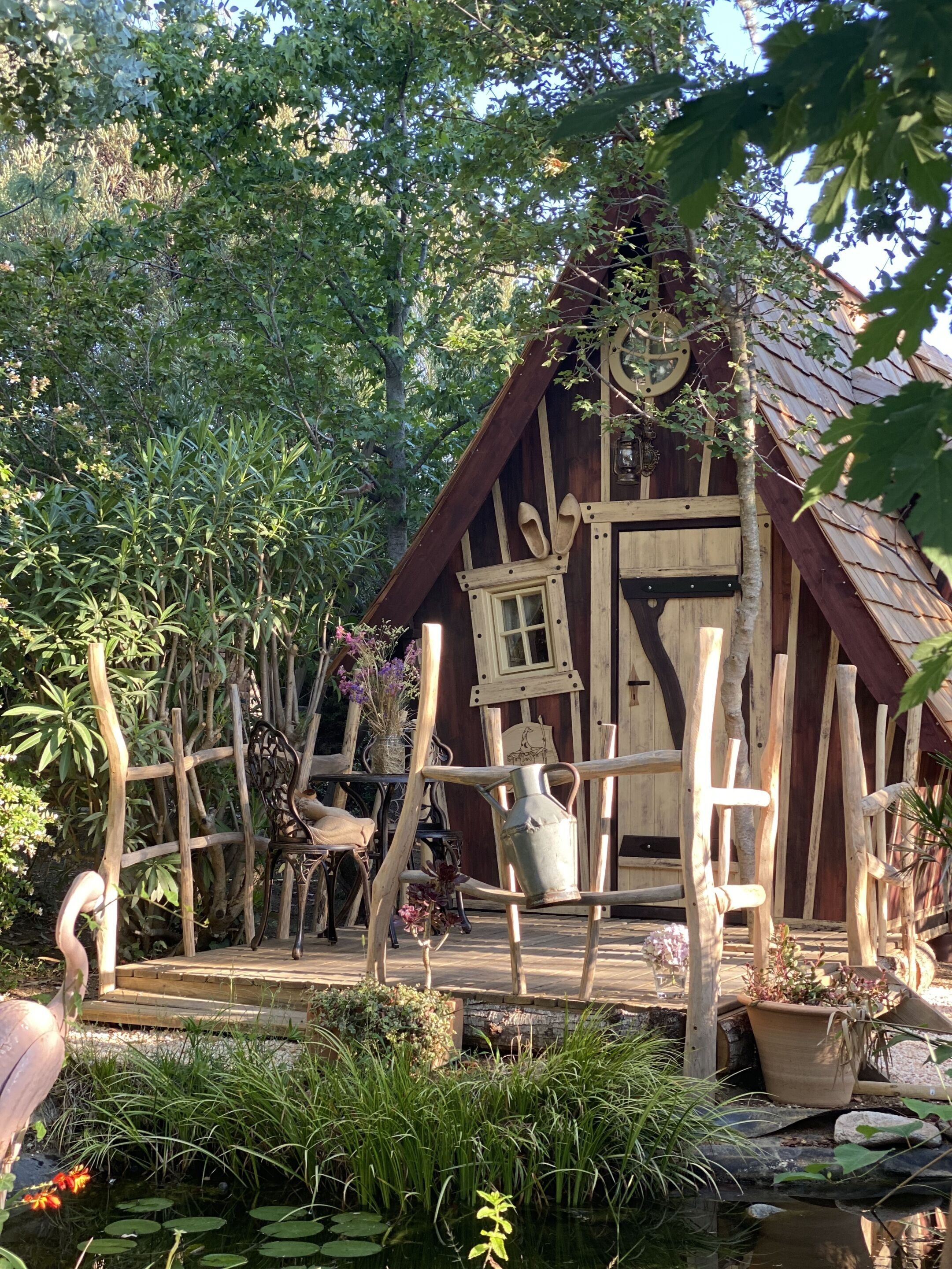 A rustic wooden cabin with a small window and door, surrounded by lush greenery and a pond with lily pads.