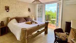 Un dormitorio con pared de piedra, estructura de cama de madera, mesita de noche, una silla y vistas a la vegetación a través de una puerta de vidrio.