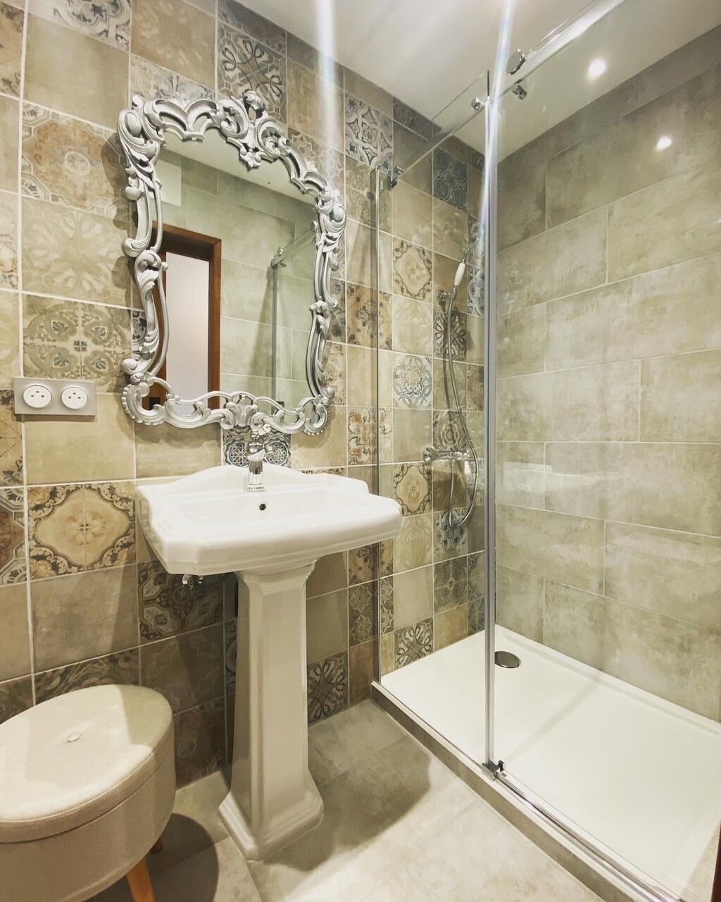 A bathroom with a pedestal sink, a large ornate mirror, a shower enclosure, and patterned tiles.
