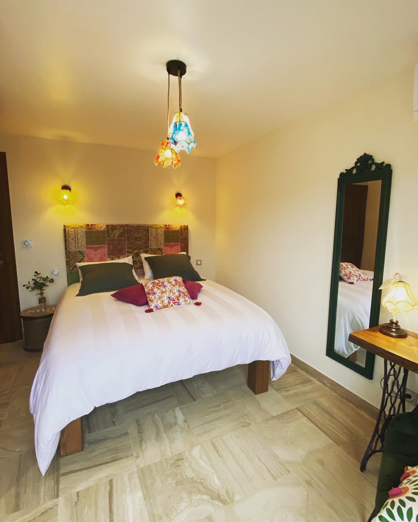 Una cama bien hecha con edredón blanco y almohadas decorativas, una lámpara colgante, un espejo y una mesita de noche con lámpara.
