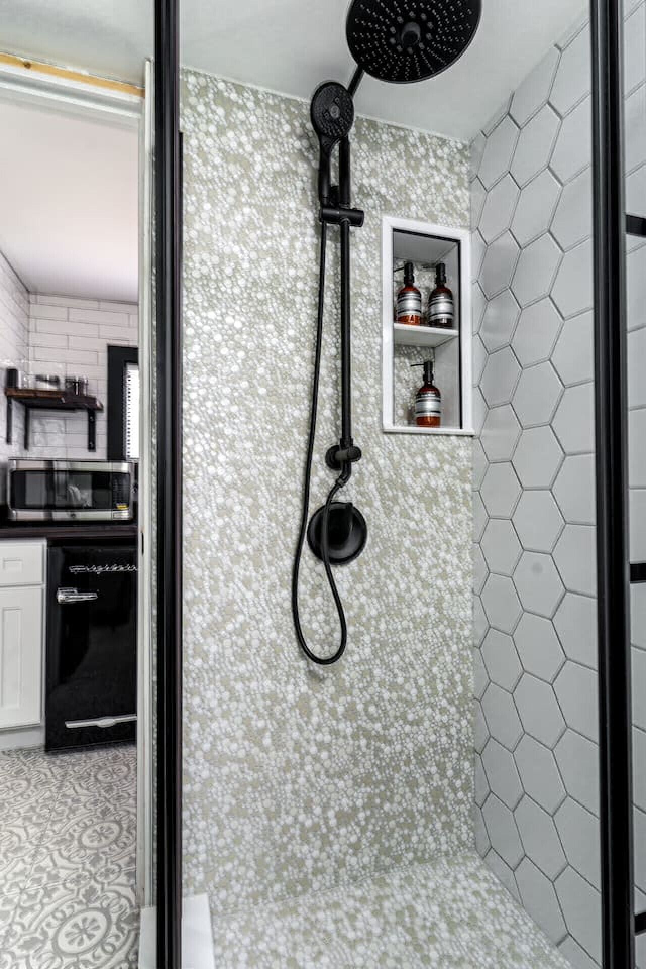 A modern bathroom with a shower, a small shelf with toiletries, and a kitchen area visible through an open door.