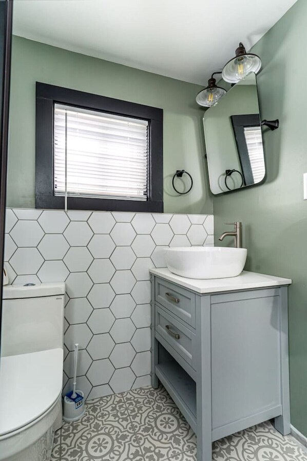 A bathroom with a white hexagonal tile backsplash, a white sink with a vessel basin, a toilet, and a window with blinds. - Cozy Cabin in a Rural setting #8 (Temple)
