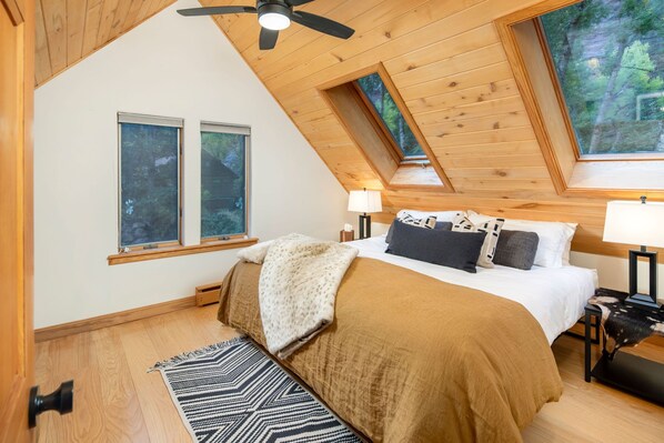 A bedroom with a sloped ceiling, a bed with a brown blanket, two bedside lamps, and a window with a view of trees. - The River House: San Miguel Sanctuary (Telluride)