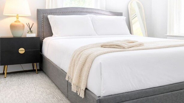 Un lit bien fait, avec du linge de lit blanc et un plaid beige, une table de chevet avec une lampe, et un miroir au mur.
