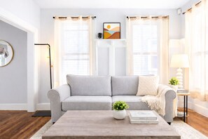 Ein Wohnzimmer mit grauem Sofa, einem Couchtisch mit Pflanze und Büchern, einer Stehlampe und einem Bild an der Wand.