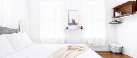 A modern bedroom with a large bed, a bedside table, a hanging light, and a window with sheer curtains.