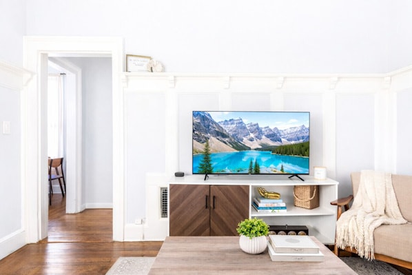 En stue med et fladskærms-tv monteret på en hvid væg, et træbord med en plante og en beige sofa.