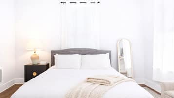 Una cama bien hecha con edredón blanco y almohadas, una lámpara de noche, un espejo y una mesita de madera.
