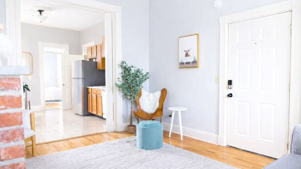 Un salon moderne avec un canapé gris, une petite table ronde et une chaise bleue. On y trouve une porte blanche et un tableau encadré accroché au mur.
