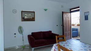 A room with a red sofa, a wooden chair, a table with a floral tablecloth, a wall clock, a framed picture, and a door leading outside. - SEA HOUSE KAMARATON (Marina di Camerota)
