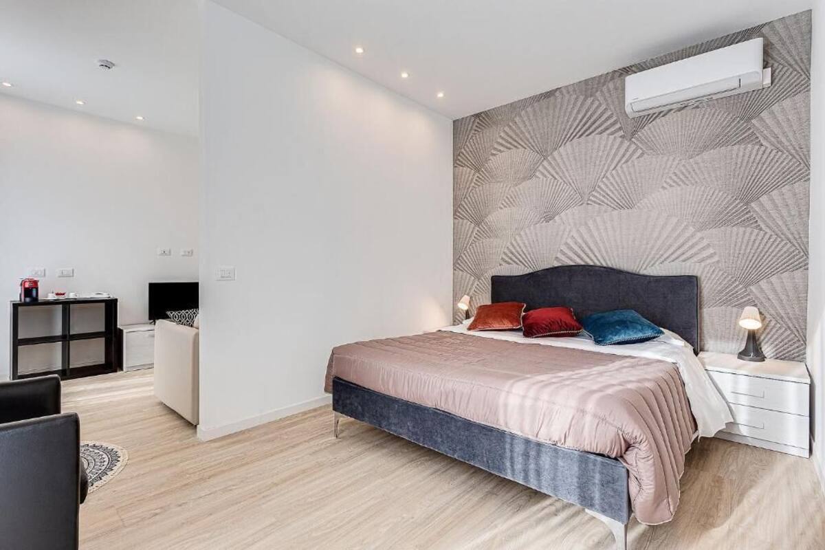 A modern bedroom with a large bed, a headboard with a geometric pattern, bedside tables with lamps, and a small desk with a chair.
