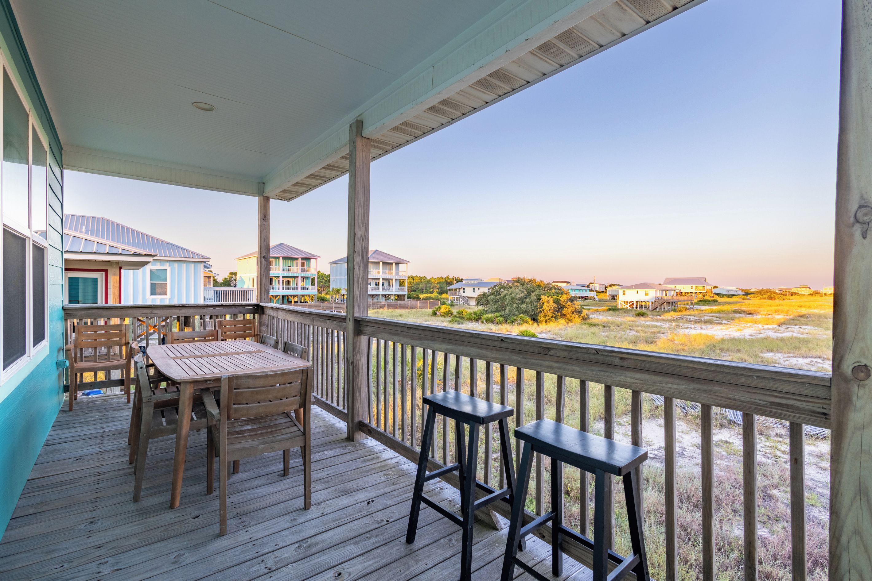 A wooden deck with a table and chairs, overlooking a beach with houses in the background.