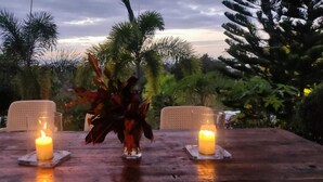 An outdoor dining table with two candles and a centerpiece, surrounded by lush greenery.
