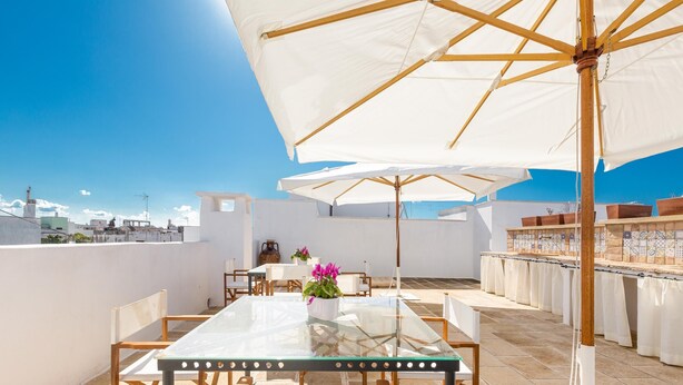 Una terraza en la azotea con una mesa de vidrio, sillas blancas y sombrillas grandes.