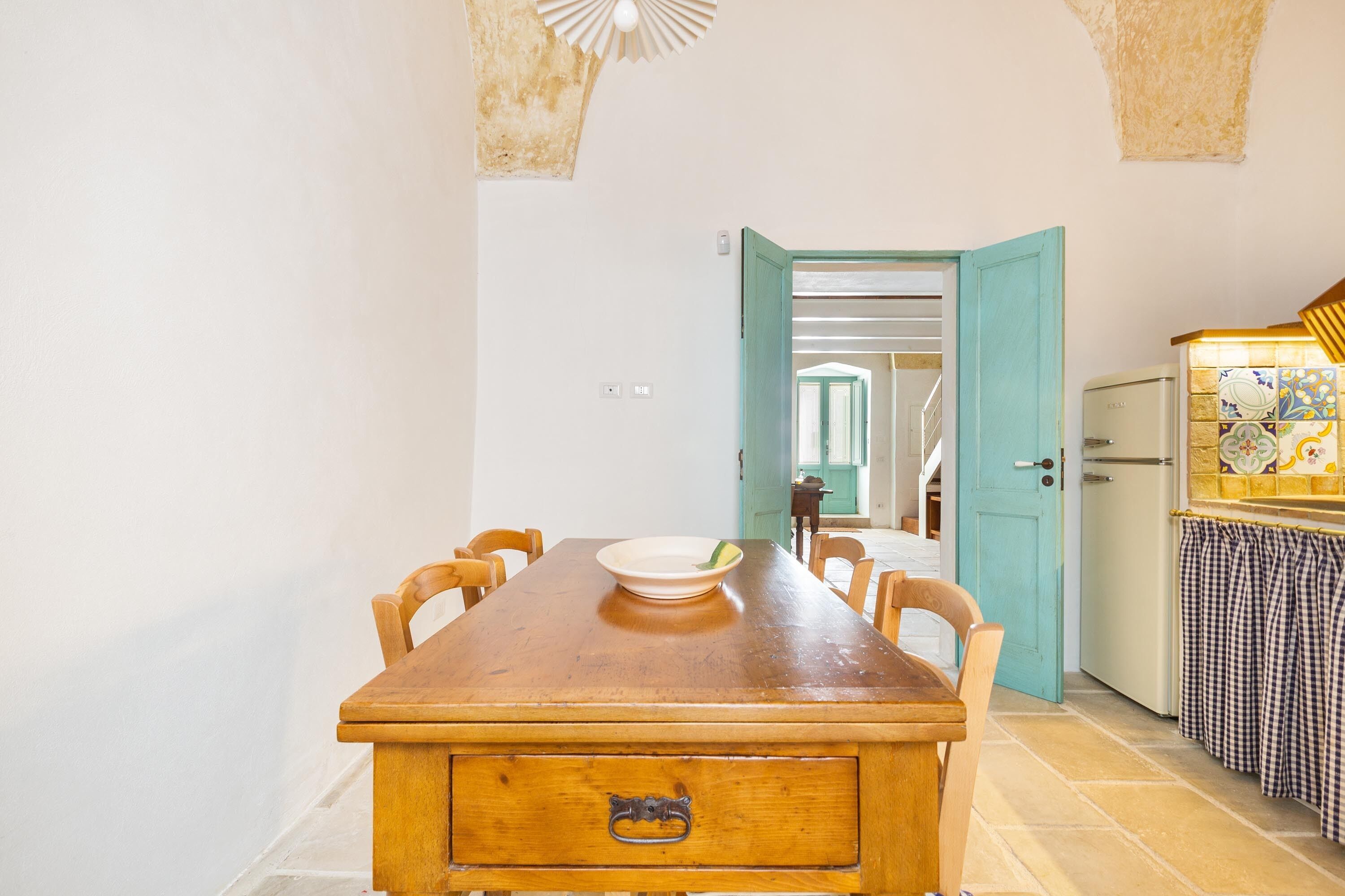 A dining area with a wooden table and chairs, a bowl on the table, and a kitchen area with a refrigerator and tiled backsplash.