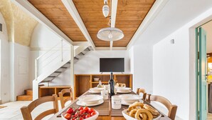 Un comedor con una mesa de madera dispuesta para una comida, rodeada de sillas. Un televisor está montado en la pared sobre la mesa.