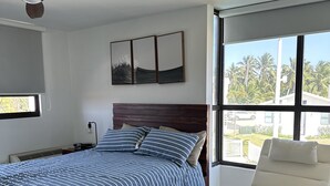 Ein Schlafzimmer mit einem Bett, einem Deckenventilator, einem Fenster mit Blick auf Palmen und einem weißen Stuhl.