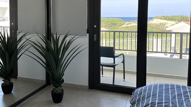 Un dormitorio con una cama grande, un ventanal corredero que da a un balcón con vista al mar y vegetación.