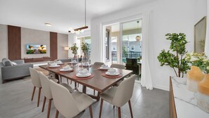 Ein moderner Essbereich mit einem Holztisch, gedeckt für sechs Personen, umgeben von Stühlen. Im Hintergrund befindet sich eine Couch und ein Fernseher.