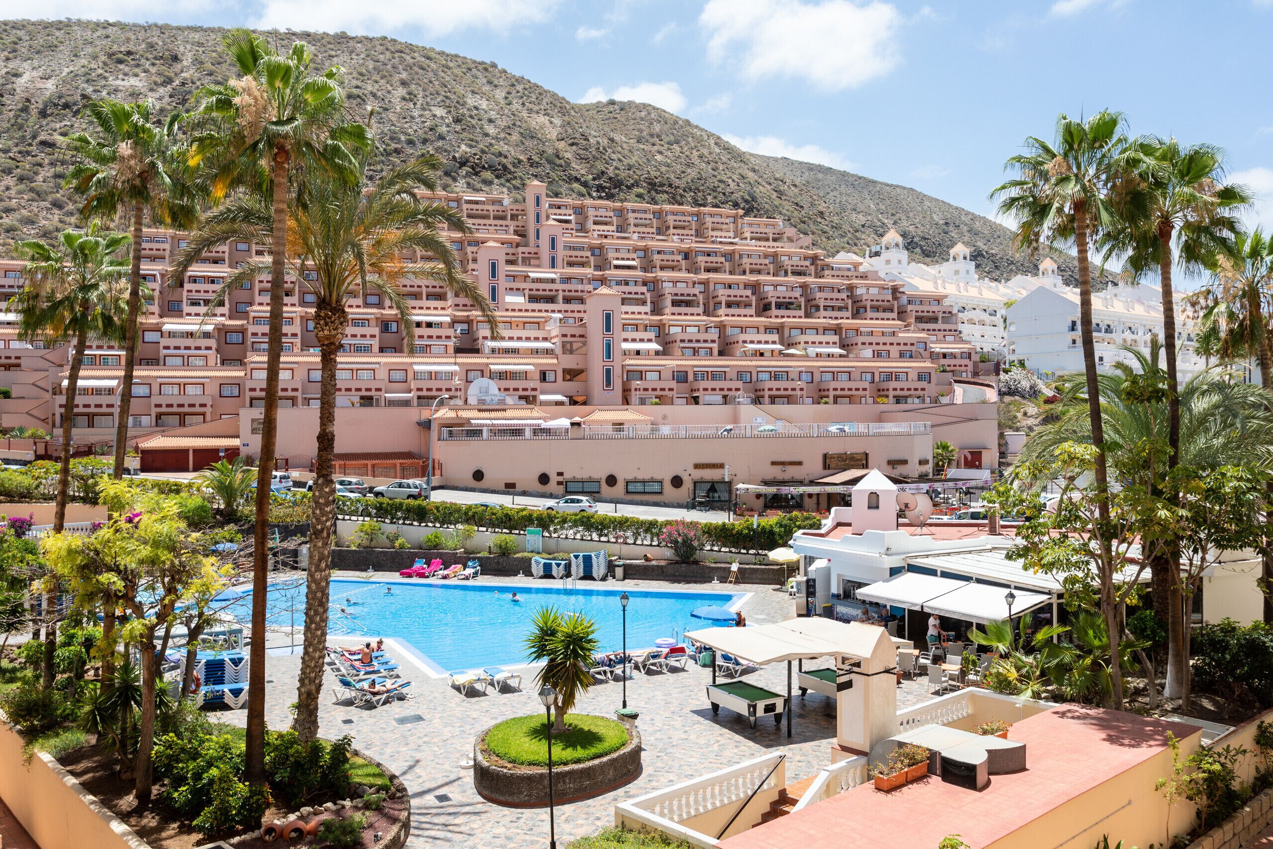 A resort with a swimming pool, palm trees, and a mountainous backdrop.