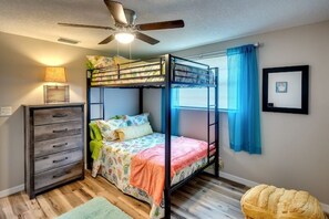 A bunk bed room with a ceiling fan, a dresser, a window with blue curtains, and a framed picture on the wall.