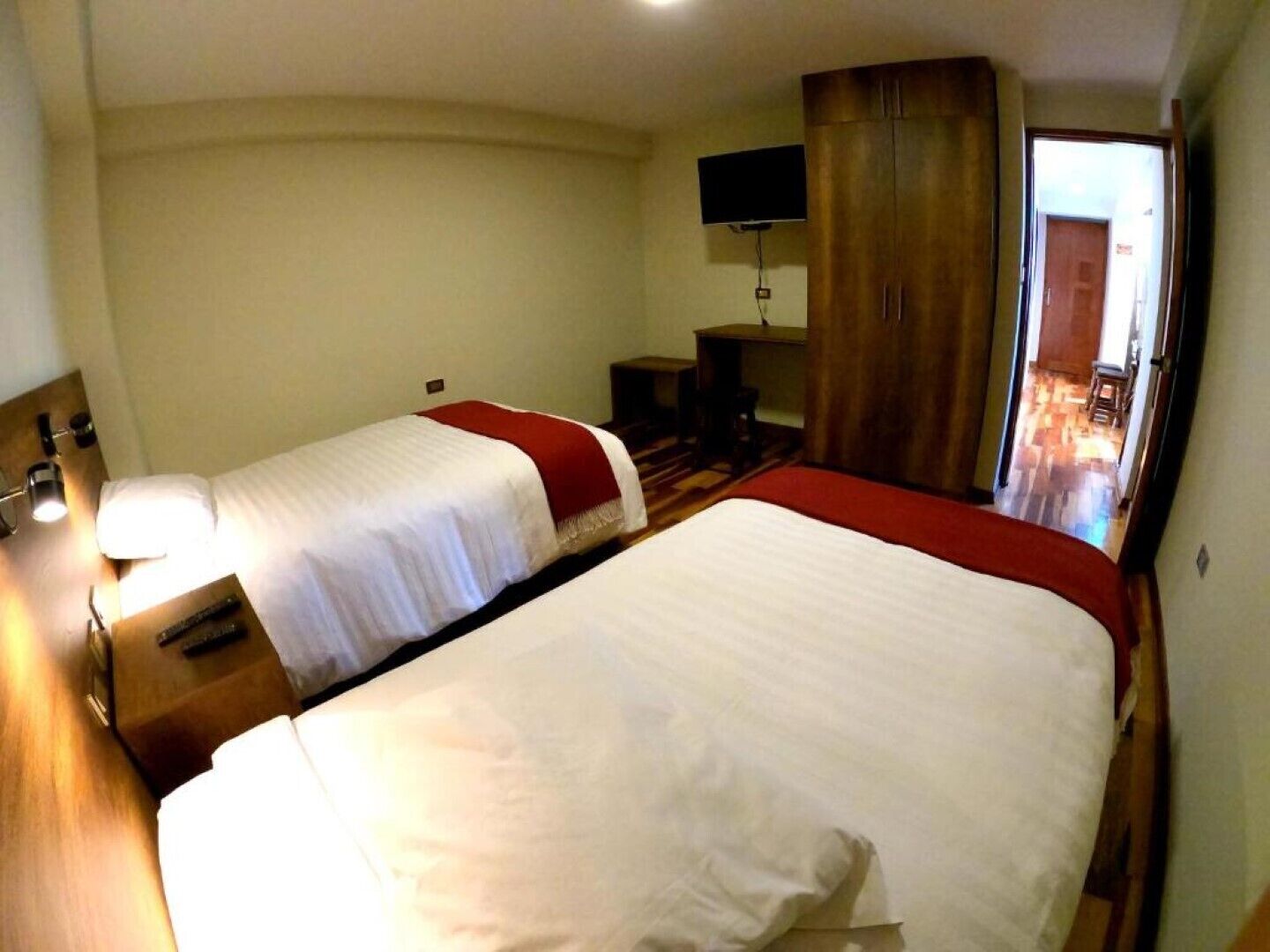 A hotel room with two beds, a TV mounted on the wall, a wooden wardrobe, and a desk with a chair.