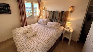 A neatly made bed with a checkered bedspread, a bedside table with a lamp, and a view of the sea through a window with curtains. - Balcon des îles d'Or - T2 seafront historic center (Le Lavandou)