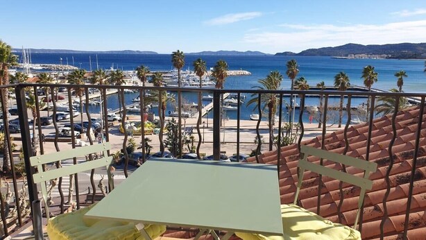 A balcony with a table and chairs, overlooking a marina with boats and palm trees, and a view of the sea and distant hills. - Balcon des îles d'Or - T2 seafront historic center (Le Lavandou)