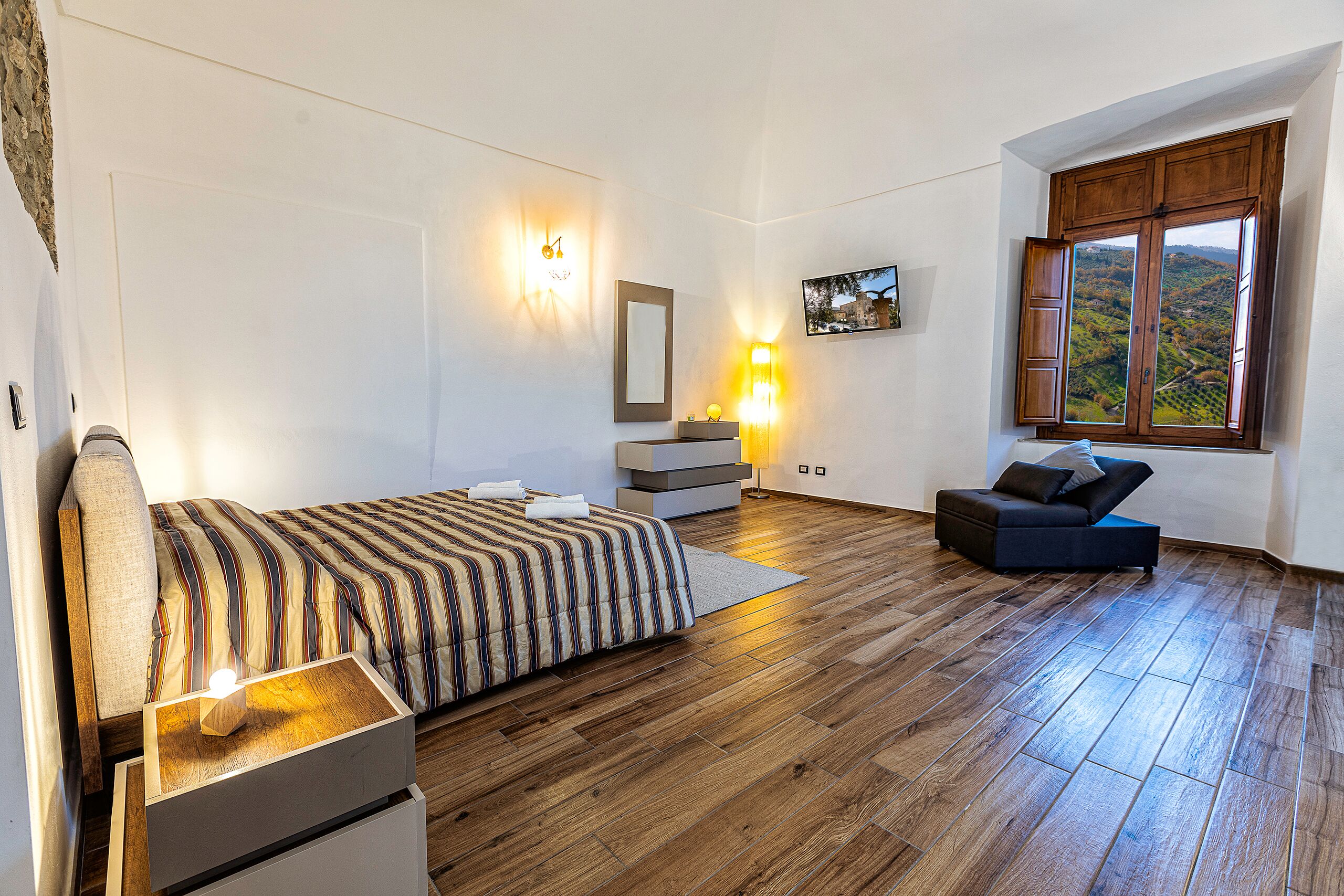 Una camera da letto con un letto, un comodino, una lampada e vista su un paesaggio montuoso attraverso una finestra.