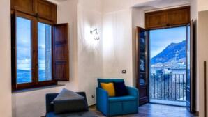 Amplia sala de estar con piso de madera, un sofá azul y un reposapiés negro. Dos ventanas grandes ofrecen vistas a una montaña y un pueblo.
