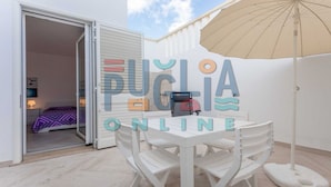 Un patio exterior con una mesa y sillas blancas, un paraguas color beige, y la entrada a una habitación con una cama y una mesita de noche.