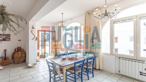 A dining area with a table set for four, blue chairs, a chandelier, and a view of the street through large windows. - Villa Pisa 1 in Torre Vado (torre vado)