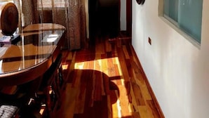 Um corredor com piso de madeira, uma mesa de jantar com cadeiras, um abajur e uma janela grande com cortinas.